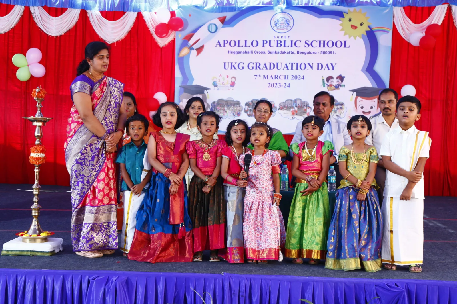 Young students performing on a decorated stage with a lit ceremonial lamp