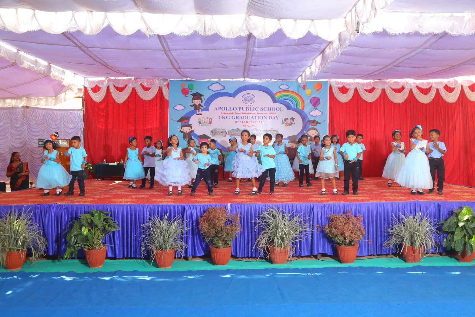 A teacher with UKG children in blue outfits on a decorated graduation stage